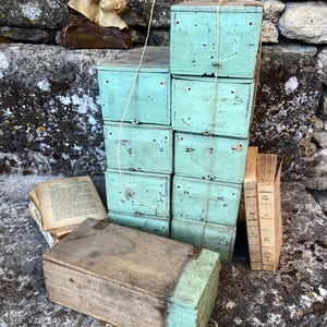 Puede incluir: Una pila de ocho cajas rectangulares de madera pintadas con un acabado verde claro desgastado, atadas con cordel fino. Una caja rectangular de madera con tapa de madera natural y base verde se sitúa delante, junto a dos libros antiguos.