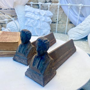 Puede incluir: Dos sujetalibros antiguos de hierro fundido, cada uno con un busto de una persona. Los sujetalibros son de color marrón oscuro con una base rectangular y un soporte largo y plano. Un libro antiguo está apilado sobre papel envejecido en una mesa blanca.