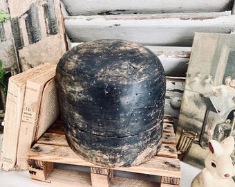May include: A large, weathered, dark-coloured hat form sits on a small wooden pallet. The hat form is round with a split in the middle. Beside it are old books and a vintage painting.