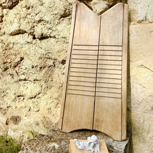 Puede incluir: Un antiguo lavadero de madera con ranuras horizontales, apoyado contra una pared de piedra. Una pequeña estatua blanca de un ángel descansa sobre un libro frente al lavadero. El lavadero es de madera clara.