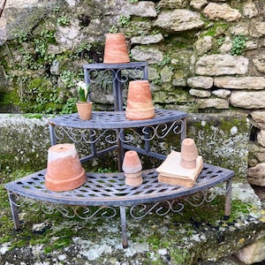 Könnte beinhalten: Ein mehrstufiger Pflanzständer aus Schmiedeeisen zeigt mehrere Terrakottatöpfe unterschiedlicher Größe. Eine kleine Topfpflanze und ein Stapel gealterten Papiers sind ebenfalls vorhanden. Der Ständer steht vor einer Steinmauer mit Moos und Grün.