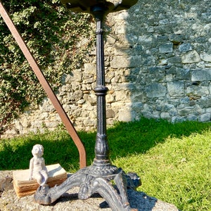 May include: A black cast iron bistro table base with a round top and ornate legs. The table base is set on a stone surface with a small statue and a stack of old books nearby. The background features a stone wall and greenery.