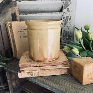 Peut inclure: Un pot en céramique vintage de couleur crème avec une finition rustique et glacée. Le pot est posé sur une pile de vieux livres avec une barre de savon au premier plan.