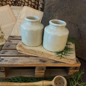 Puede incluir: Dos jarras de cerámica blanquecina, con cuerpos redondeados y bocas abiertas, descansan sobre papel envejecido. Las jarras se exhiben en una pequeña paleta de madera, con cucharas de madera y vegetación. Un cesto tejido y un trozo de madera completan la escena.