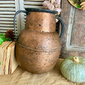 May include: A large copper pitcher with two black iron handles. The pitcher has a rustic, aged look and is sitting on a burlap surface.