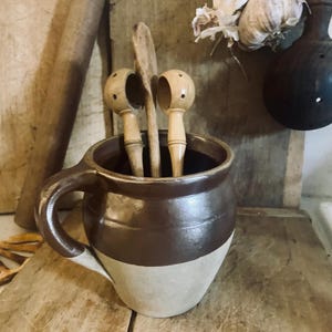 May include: A brown and beige ceramic pitcher with a handle, holding wooden kitchen utensils. Two wooden nutcrackers and a wooden spoon are inside the pitcher. The pitcher is on a wooden surface, with a rolling pin and garlic in the background.