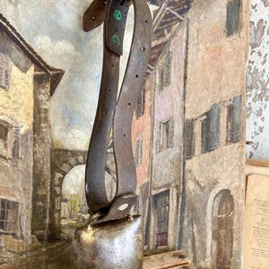 Może przedstawiać: Duży, antyczny dzwonek krowy z wytartym, ciemnoszarym wykończeniem i skórzanym paskiem z mosiężnymi okuciami. Dzwonek jest zawieszony na drewnianej powierzchni.