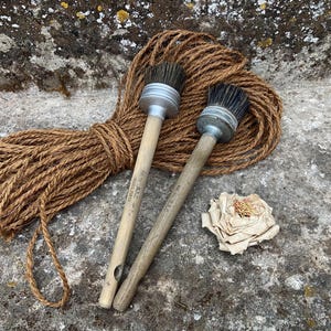 Op de afbeelding: Twee ronde verfkwasten met houten stelen en zilverkleurige metalen hulzen met donkere haren, geplaatst naast een opgerold stuk bruin jutekoord en een enkele decoratieve papieren bloem met gelaagde bloemblaadjes en een lichtbruin hart.