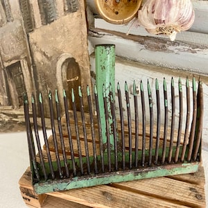 Puede incluir: Una antigua herramienta de jardín pintada de verde con múltiples púas. La herramienta es de metal y tiene un aspecto desgastado. Las púas son de color marrón oscuro con puntas verdes. La herramienta está sobre un pequeño palet de madera.