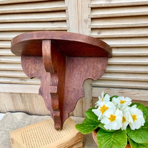 Puede incluir: Un estante de esquina de madera oscura con un borde festoneado y una parte superior redondeada. El estante está colocado sobre una pila de libros y una maceta blanca con flores blancas.