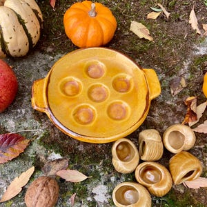 May include: A yellow ceramic escargot dish with seven indentations, accompanied by several snail-shaped serving dishes. The image also features a small orange pumpkin, a gourd, a walnut, and autumn leaves.