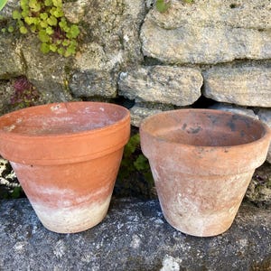 Puede incluir: Dos macetas de terracota de diferentes tamaños se asientan sobre una superficie de piedra frente a una pared de piedra. Las macetas son de color marrón rojizo con depósitos minerales blancos cerca de la base. Las macetas están vacías.