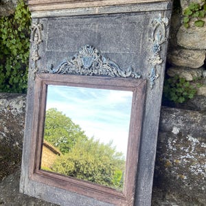 Puede incluir: Un espejo rectangular antiguo con un marco gris desgastado y un marco interior marrón. El espejo tiene elementos decorativos ornamentados, azul claro y crema en la parte superior y los lados. El espejo refleja una vista de árboles y cielo.