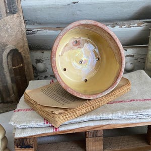 Op de afbeelding: Een vintage, ronde, gele en bruine keramische plantenbak met drainagegaten. De plantenbak staat op een stapel oude boeken en een linnen doek met een rode streep. De achtergrond bevat antieke kunst en een decoratief konijn.