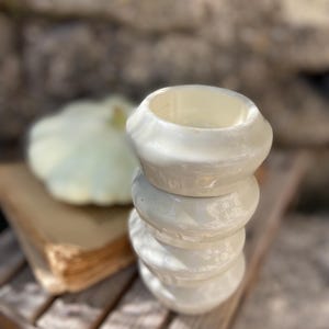 Pode incluir: Uma pilha de quatro taças de cerâmica em tom branco sujo com um acabamento brilhante e iridescente. As taças são circulares com um perfil arredondado e bojudo, criando uma forma cilíndrica em camadas. A superfície reflete a luz.