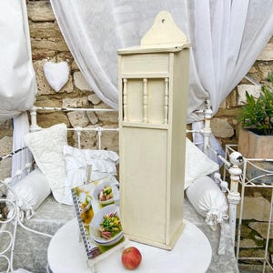 Può includere: Un mobile alto in legno bianco sporco con una parte superiore decorativa e barre verticali. Il mobile è posto su un tavolo bianco, con un libro di cucina e una mela nelle vicinanze. Lo sfondo include un letto bianco e un muro di pietra.
