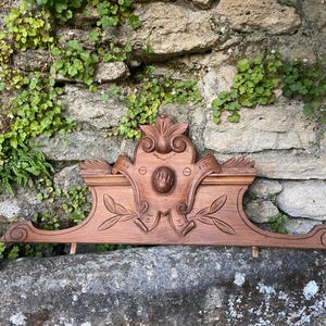 Può includere: Un elemento architettonico in legno intagliato con dettagli ornamentali, tra cui foglie e un medaglione centrale. Il pezzo è di colore marrone caldo e sembra vintage. Una piccola statuina di coniglio in ceramica è a sinistra.