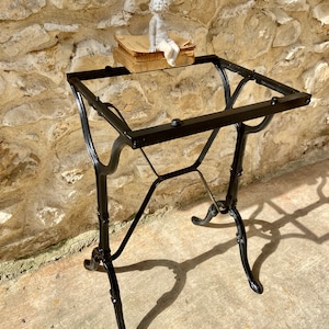 Puede incluir: Mesa de metal negro con patas ornamentadas y una parte superior rectangular. Una pequeña estatua de querubín blanco se encuentra sobre una pila de libros antiguos. La mesa se exhibe al aire libre contra una pared de piedra.