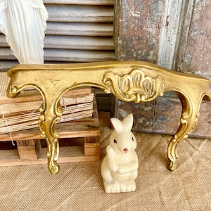 Op de afbeelding: Een gouden decoratief rek met sierlijke kruldetails. Het rek is gebogen en heeft een decoratief ontwerp aan de voorkant. Het rek staat op een houten oppervlak met een wit konijnenbeeldje ervoor.