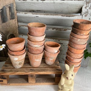 May include: A collection of small, weathered terracotta pots stacked on a wooden pallet. The pots vary in size and show signs of age, with a rustic aesthetic. A decorative bunny figurine is in the foreground.