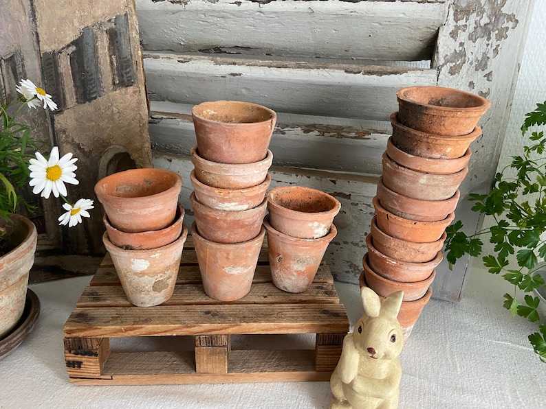 May include: A collection of small, weathered terracotta pots stacked on a wooden pallet. The pots vary in size and show signs of age, with a rustic aesthetic. A decorative bunny figurine is in the foreground.