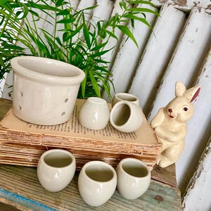 May include: A set of six small, white ceramic egg cups with a glazed finish. The cups are arranged on a stack of old books, along with a larger, white ceramic pot with drainage holes and a ceramic bunny figurine.