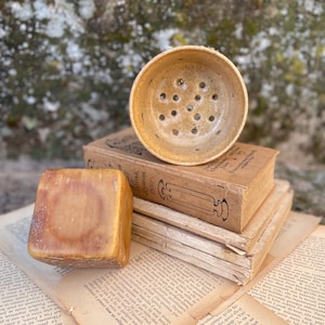 May include: A tan ceramic soap dish with a circular perforated base sits atop a stack of three vintage books with aged, yellowed pages. Beside the books rests a solid, square-shaped block of translucent amber-colored soap.