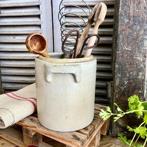 May include: A white ceramic crock with a handle, filled with wooden kitchen utensils, including a whisk, a spoon, and a pair of tongs. The crock is sitting on a small wooden pallet.