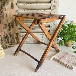 Può includere: Uno sgabello pieghevole in legno vintage con seduta a doghe. Lo sgabello è in legno marrone chiaro e ha un design delle gambe a forma di X. Sono visibili anche una piccola statuina di coniglio in ceramica e un asciugamano di lino piegato con strisce rosse.