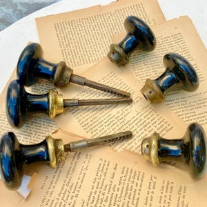 May include: A collection of six antique black door knobs with brass accents and long, slotted stems. The knobs are arranged on aged, yellowed pages of a book, creating a vintage aesthetic.