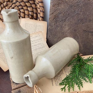 Può includere: Due antiche bottiglie in gres color beige con beccuccio e base arrotondata. Una bottiglia è in posizione verticale, l'altra è sdraiata. Le bottiglie poggiano su carta e legno invecchiati, con un cesto intrecciato e del verde sullo sfondo.