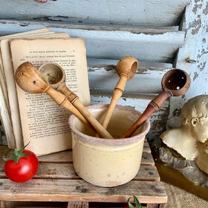 May include: Four wooden spoons with holes in the bowls are in a beige ceramic crock. The crock is sitting on a wooden pallet with red tomatoes scattered around it.