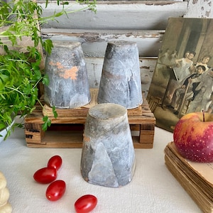 Op de afbeelding: Drie grijze, kegelvormige objecten met een rustieke uitstraling worden tentoongesteld op een klein houten pallet. Een rood-gele appel ligt op een stapel oude boeken. Een kleine keramische konijn en verschillende rode tomaten zijn ook aanwezig.