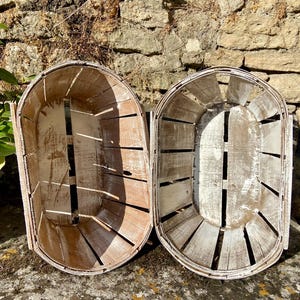 May include: Two oval-shaped, wooden baskets with a distressed, whitewashed finish. The baskets have a slatted design and are set against a stone wall. One basket has a warmer, brown tone, while the other is lighter.