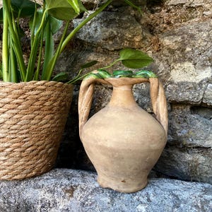 May include: A terracotta amphora with a rounded body, a narrow neck, and two thick, twisted handles. The rim of the vessel features a glossy, dark green glazed finish that contrasts with the matte, earthy brown texture of the clay body.