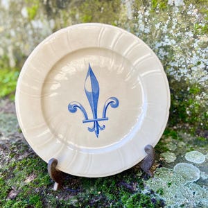 Könnte beinhalten: Ein cremefarbener Teller mit blauem Fleur-de-Lis-Design. Der Teller hat einen gewellten Rand und wird auf einem dekorativen Metallständer präsentiert. Der Teller ist ein dekorativer Gegenstand.