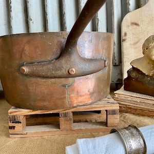 Puede incluir: Una olla grande de cobre con un asa resistente. La olla está sentada sobre una plataforma de madera y tiene un aspecto rústico y vintage.