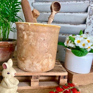 Op de afbeelding: Een rustieke bruine keramische pot met een gele glazuur, gevuld met houten keukengerei, waaronder een deegroller en een geperforeerde lepel. De pot staat op een houten pallet, naast een witte keramische pot met witte bloemen en een klein keramiek konijnenbeeldje. Een takje rode cherrytomaten bevindt zich op de voorgrond.