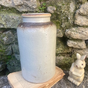 May include: A large, cylindrical, light grey stoneware jar with a brown rim. A small, cream-coloured ceramic bunny figurine sits to the right. The jar rests on a piece of aged paper, set against a stone wall.