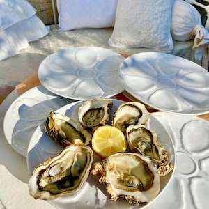 Puede incluir: Un plato de ostras frescas con una rodaja de limón, servido en un plato de ostras blanco. Se ven varios otros platos de ostras blancos, junto con almohadas blancas y una silla de metal blanco.