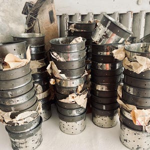 Op de afbeelding: Een verzameling vintage metalen kaasvormen, gestapeld in meerdere stapels. De vormen zijn cilindrisch met geperforeerde zijkanten en een zilver-grijze afwerking. Sommige zijn verpakt met verouderd papier.