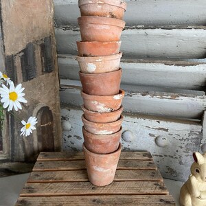 May include: A stack of eleven small, weathered terracotta flower pots. The pots are stacked on a wooden pallet, with a painting and a decorative bunny in the background. The pots have a rustic, aged appearance, with visible wear and tear.
