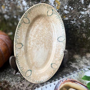 Könnte beinhalten: Ovale Servierplatte im antiken Stil mit einer gesprenkelten beigefarbenen Oberfläche. Der Rand ist mit einem grün-weißen Muster verziert. Die Platte wird auf einem dunklen Metallständer präsentiert, was eine rustikale Ästhetik suggeriert.
