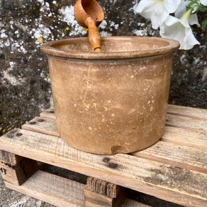 May include: A brown, speckled ceramic pot with a wooden ladle resting inside. The pot sits on a wooden pallet. White flowers are in the background. The pot has a diameter of approximately 8 inches.