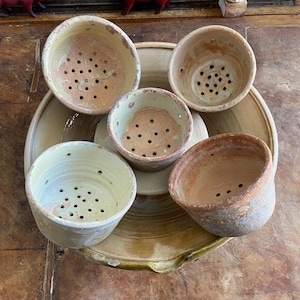 May include: A collection of five small, round, terracotta-colored ceramic bowls with perforated bases, arranged inside a larger, shallow, circular ceramic basin. The bowls feature varying shades of beige, cream, and earthy brown, with visible drainage holes.