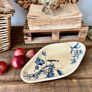 Könnte beinhalten: Eine ovale, cremefarbene Servierplatte aus Keramik mit blauem Blumendesign. Rote Äpfel sind auf einer rustikalen Holzoberfläche um die Platte angeordnet. Ein Weidenkorb und ein Stapel alter Bücher befinden sich im Hintergrund.