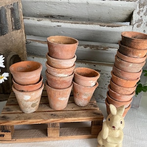 May include: A collection of small, weathered terracotta pots stacked on a wooden pallet and arranged on a white surface. The pots vary in size and show signs of age, with a few daisy flowers and a decorative bunny figurine nearby. The scene evokes a rustic, vintage aesthetic.