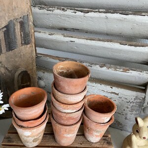 May include: A stack of small, terracotta-colored flower pots. The pots are stacked on a wooden pallet, with a weathered, rustic appearance. The background includes a white-painted shutter and a decorative bunny figurine.