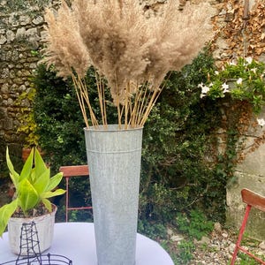 Może przedstawiać: Wysoki, ocynkowany metalowy wazon wypełniony suszoną trawą pampasową. Wazon jest cylindryczny i stoi na białym obrusie. Tło obejmuje kamienny mur, zieleń i mały stolik z rośliną doniczkową.