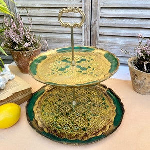 Puede incluir: Bandeja de servir de dos niveles con un diseño dorado y verde. El nivel superior tiene un patrón floral y el nivel inferior tiene un patrón geométrico. La bandeja tiene un asa de metal.